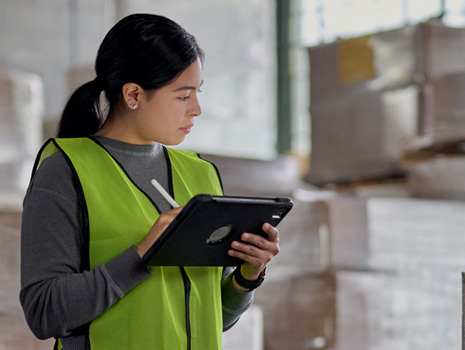 Mulher numa fábrica com um iPad na mão a controlar o inventário. Atrás dela, há várias pilhas de caixas grandes.