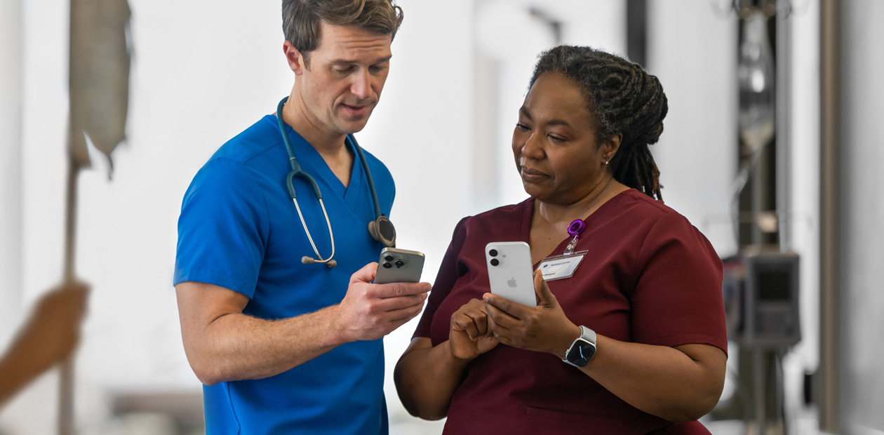 Um profissional da saúde de uniforme azul olha para seu iPhone e mostra algo nele para uma profissional de uniforme vermelho, que também tem um iPhone.