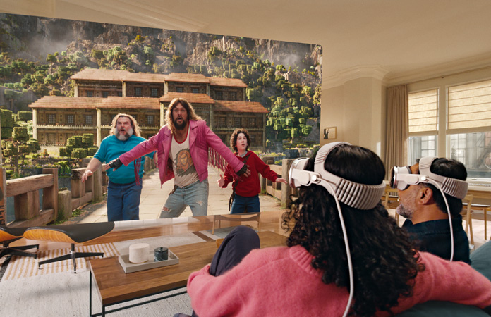 شخصان يستخدمان Apple Vision Pro، وهما جالسان جنباً إلى جنب على أريكة ويشاهدان فيلم A Minecraft Movie يُعرض على شاشة تمتد على طول الغرفة