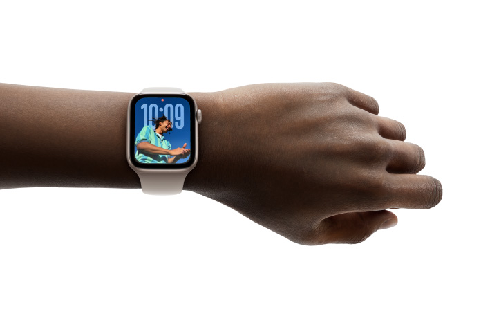 Pohled zblízka na stříbrné Apple Watch s gumovým řemínkem nasazené na zápěstí, je vidět aplikace Hodiny v novém Liquid Glass designu